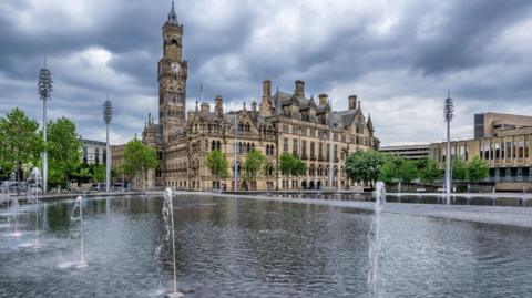 Bradford Town Hall stands behind a large pool of water with several jets of water shooting up from the pool.