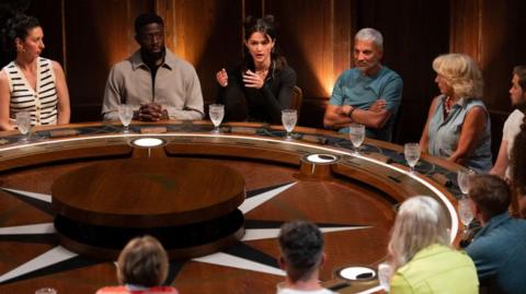 Maz, who is wearing a blue T-shirt, is sitting at the large wooden roundtable alongside the other contestants during a banishment. He has his arms folded and is staring straight ahead. 