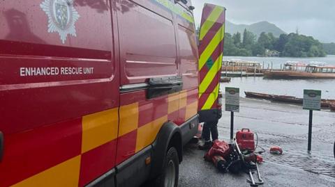 A red emergency rescue unit is parked beside a pier at Derwent Water. There are other boats tied up to the pier with the Lake District fells in the background.