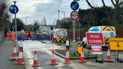 A road with two Thames Water vans parked behind a line of traffic cones with a group of engineers wearing high-viz overalls in the background