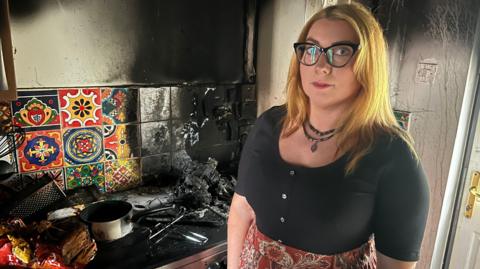 Jessica Drake stands looking at the camera. She has blonde hair and black rimmed glasses stands. Behind her is the blackened remains of a burnt out kitchen.