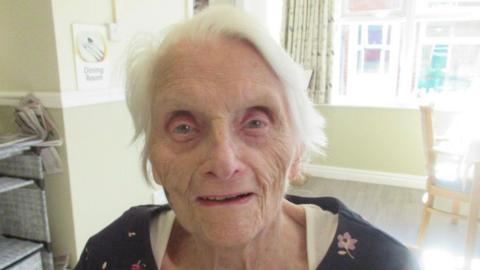 An elderly woman smiling at the camera. She has grey hair and is wearing a white top with a black top over it with pink small flowers. She is inside a living room with chairs and shelves in the background.