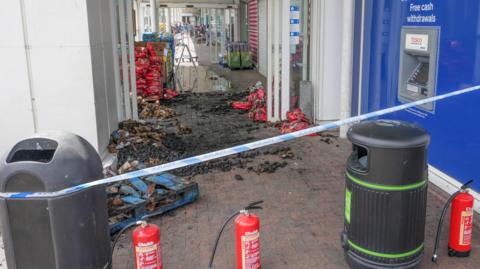 A Tesco store with police tape around it. Burned charcoal sits in the doorway to the shop.