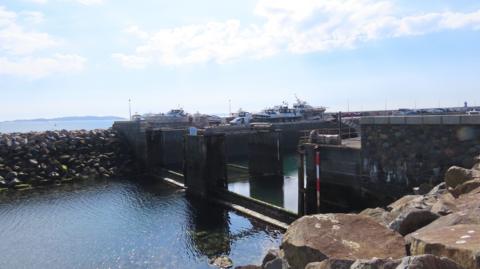 The QEII Marina's gates. Water is low and there is rock armour in the foreground and some boats in the distance. 