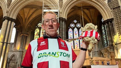 Stuart Boyfield wearing a Lincoln City red and white shirt and holding a model of the imp wearing a shirt. He has short dark hair and is wearing glasses. 