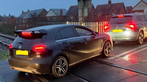 A black car idle on Willington level crossing behind another idle car.