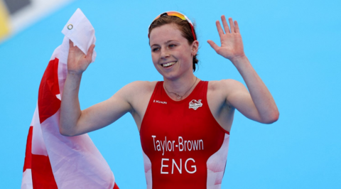 The image shows an athlete dressed in a red sports outfit with the name "Taylor-Brown ENG" printed on the front, indicating they represent England. The athlete is holding a red and white flag. The background is blue.