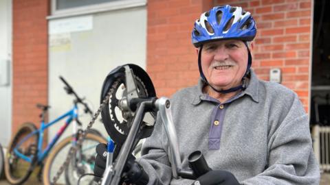 Alan is wearing a helmet and gloves, adjusting the handlebars of a red adapted tricycle outside a brick building, with several bicycles parked nearby