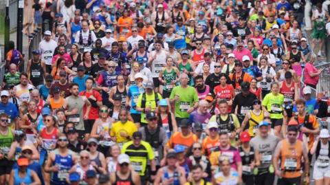Marathon runners fill the entire frames as they run towards the camera as part of the London Marathon. they are wearing a variety of coloured shirts. The crowd includes men and women.