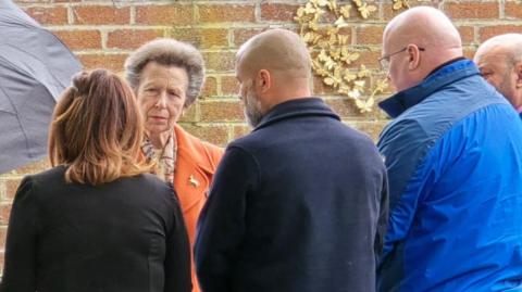 Princess Anne is wearing an orange coat and is talking to four people in a garden in Speke