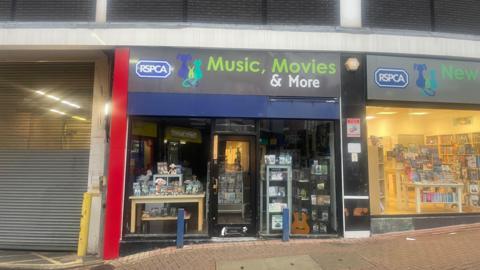 The RSPCA's Music, Movies and More shop which has its lights switched off. There is a large loading bay with shutters to the left hand side and another charity shop to the right hand side. 