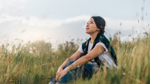 A woman is sat in a field of long grass looking at the sky. She has a white T-shirt on and a blue jumper over her shoulders with long dark hair.