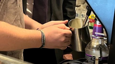 A barista prepares steamed milk - they have two twine bands around their wrist and are holding a steel jug under a steam spout on a coffee machine.