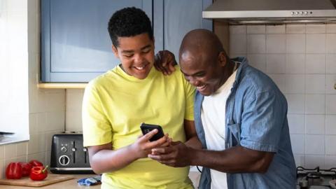 A smiling teenager in a yellow t-shirt shows his smiling father in a blue denim jacket and white t-shirt something on his phone in a kitchen 
