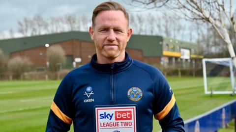 Shrewsbury Town head coach Gavin Cowan holds the League Two manager of the month award with the training ground pitch in the background