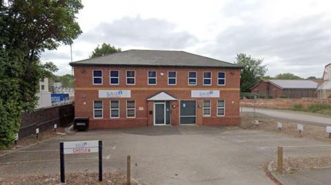 A red brick building consisting of two floors in front of an empty car park