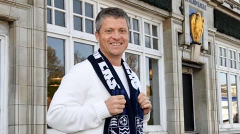 Justin Rees is wearing a white jumper with a blue Southend United scarf around his neck. He is smiling in front of the pub, which is a grey brick building with a golden statue of an eagle above its entrance door.