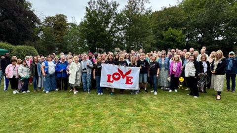 About 100 people holding a Coldplay love flag in the gardens at Holderness House. They're all smiling and grouped together.