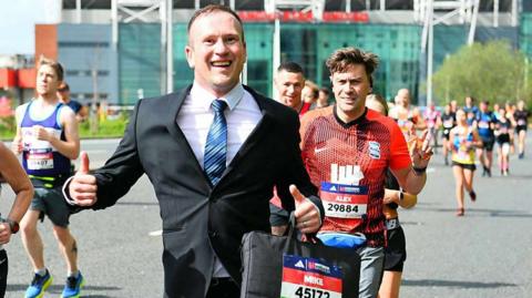 Mike Jones running in the London Marathon in 2024 wearing a black suit and carrying a briefcase, with runners behind and to the side of him.