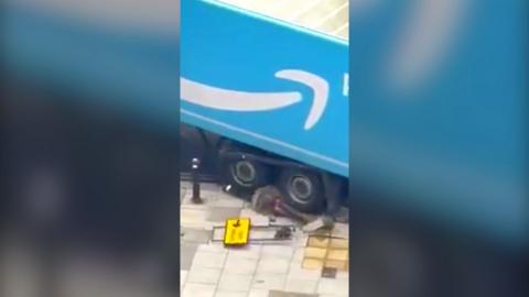 A blue Amazon branded vehicle partially mounts the pavement, pulling up paving slabs and bollards. 