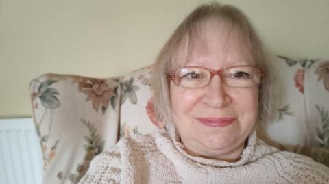 Sandra with blonde hair and wearing glasses smiles at the camera. She is sitting in a flower patterned chair in front of a radiator