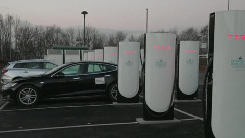 A row of charging bays in a car park, with white towers installed in every parking space. A black electric car and a white electric car are both parked up and charging.  