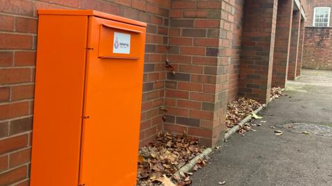 A tall orange box in front of a brick wall next to a path.