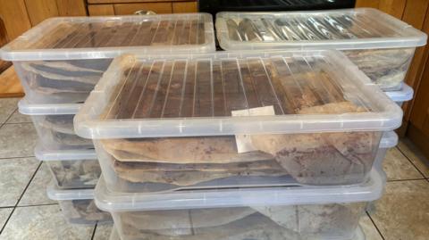 Clear large plastic boxes on a kitchen floor. Multiple are stacked on top of each other. There are three piles with about four boxes in each pile. Each boxes has brown paper stained with oil filled with brownies which you cannot see.