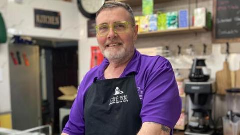 Stan Robertson, standing in a cafe, wearing a purple shirt, with a black apron, with cafe equipment behind him. He is smiling, with yellow-framed glasses on, a grey beard and short greying hair.