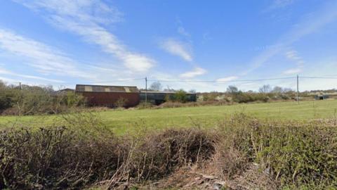 A street view image of a field with hedge in the foreground and some agricultural buildings in the background.