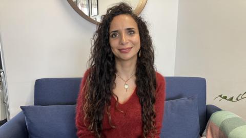 A woman with long curly dark hair sat on a blue sofa smiling. She is wearing a red wool jumper. 