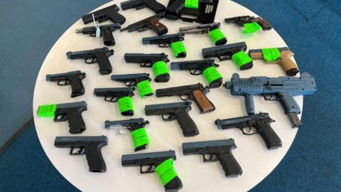 A number of guns lined up neatly on a white circle. Some have green tape around the handles.