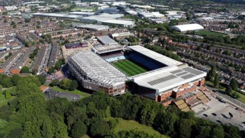 An aerial shot of a football stadium surrounded by terraced housing and other buildings in the distance as well as trees in the foreground