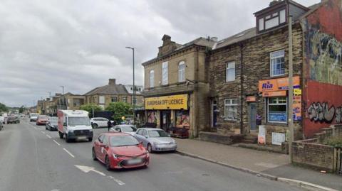 Street View image showing traffic passing by shops including one with a large sign reading European Off Licence