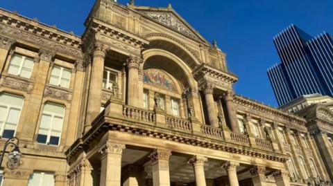 The front of a large brown building with columns and a balcony
