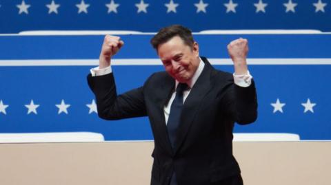 A photograph of Elon Musk in a black suit raising his fists in the air. He is standing in front of a bright blue background with white stars