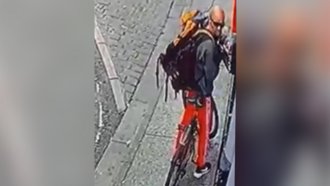 CCTV image of a man. He is white, bald, wearing sunglasses, a dark jacket, black trainers and red trousers. He is riding a bicycle. 