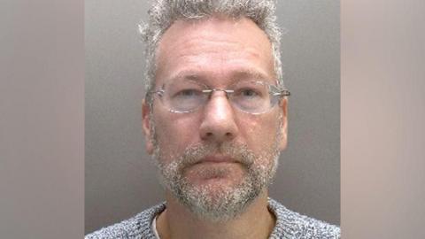 Mugshot of man with grey hair and beard and glasses