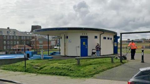 A public toilet on the West Cliff at Whitby - a small oval shaped building painted white with blue doors and small windows at roof level