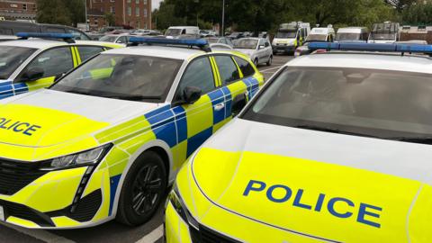 Two Wiltshire Police cars in reflective yellow stickers and the word POLICE in capitals on the bonnet. They are parked in a car park with other police vehicles.