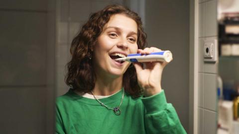A woman in a green jumper brushing her teeth with an electric toothbrush and looking in the mirror.