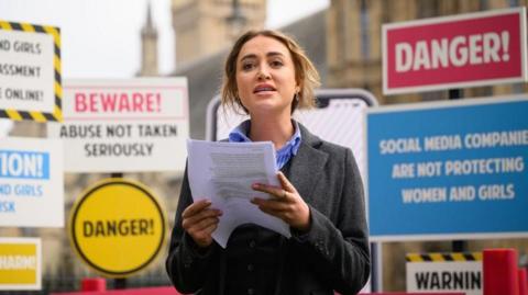 Georgia Harrison, a blonde woman wearing a blue and white stripy shirt and a grey coat, holding a piece of paper and making a speech in front of a selection of signs warning that social media companies do not take reports of abuse against women and girls seriously.
