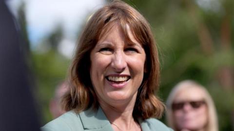 Rachel Reeves smiles while standing outside on a sunny day. She is wearing a sage green jacket, and the people and trees in the background are blurred.
