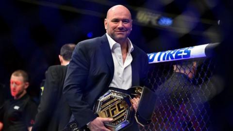 UFC boss Dana White is pictured inside the octagon holding a gold UFC belt. He is wearing a blue suit with a white shirt. 
