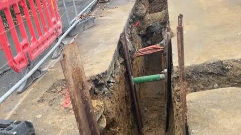 The image shows the ring main exposed with a small trench dug into a footpath. There are metal railings to the left of the main. 