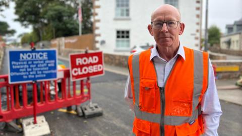A man wearing a high vis jacket with road closed and work signs in the background on a closed road. Blurred background.