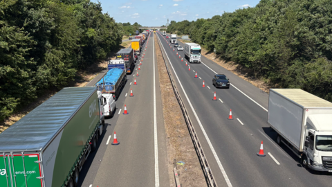 View of the M11 from a bridge over it. There are two lines of traffic, one heading away from the camera and one towards. The right lanes in both directions have been blocked off by orange traffic cones. The sky is blue and bright and there are green trees at either side of the motorway.