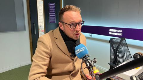 Adam Dance is pictured speaking into a blue microphone in a radio station. He is wearing a beige coat, a black scarf and round black-rimmed glasses. Around the studio is a purple banner reading BBC Radio Somerset.