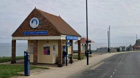 An open sided building stands next to a parking meter. There are two blue signs saying 'Lift to West Cliff beach', with 'West Cliff Whitby' above one of them, around a silhouette of a building.  There are also posters on the inner yellow walls. It is next to a road.