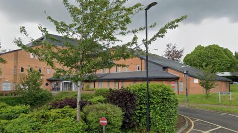 The outside of the Tewkesbury Borough Council offices - a light coloured brick building with a slanted roof. There are trees and bushes landscaped around the road that leads up to it.